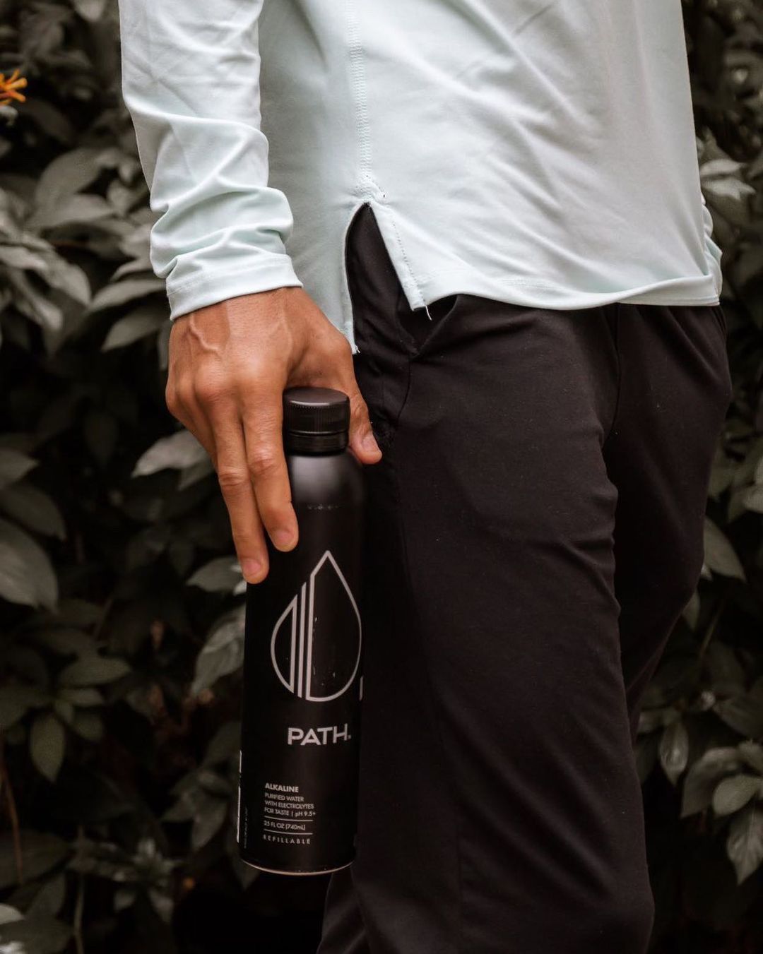 Side view of PATH water bottle showcasing matte black finish and large white PATH logo, held by a person against leafy background