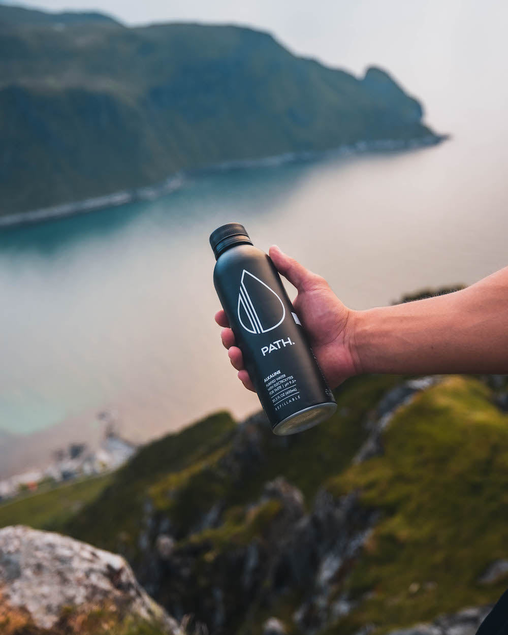 Hand holding a matte black PATH reusable water bottle above coastal cliffs and ocean at sunset
