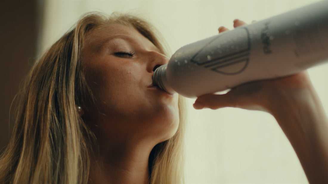 Close-up of a woman drinking from a silver metal water bottle by a sunlit window
