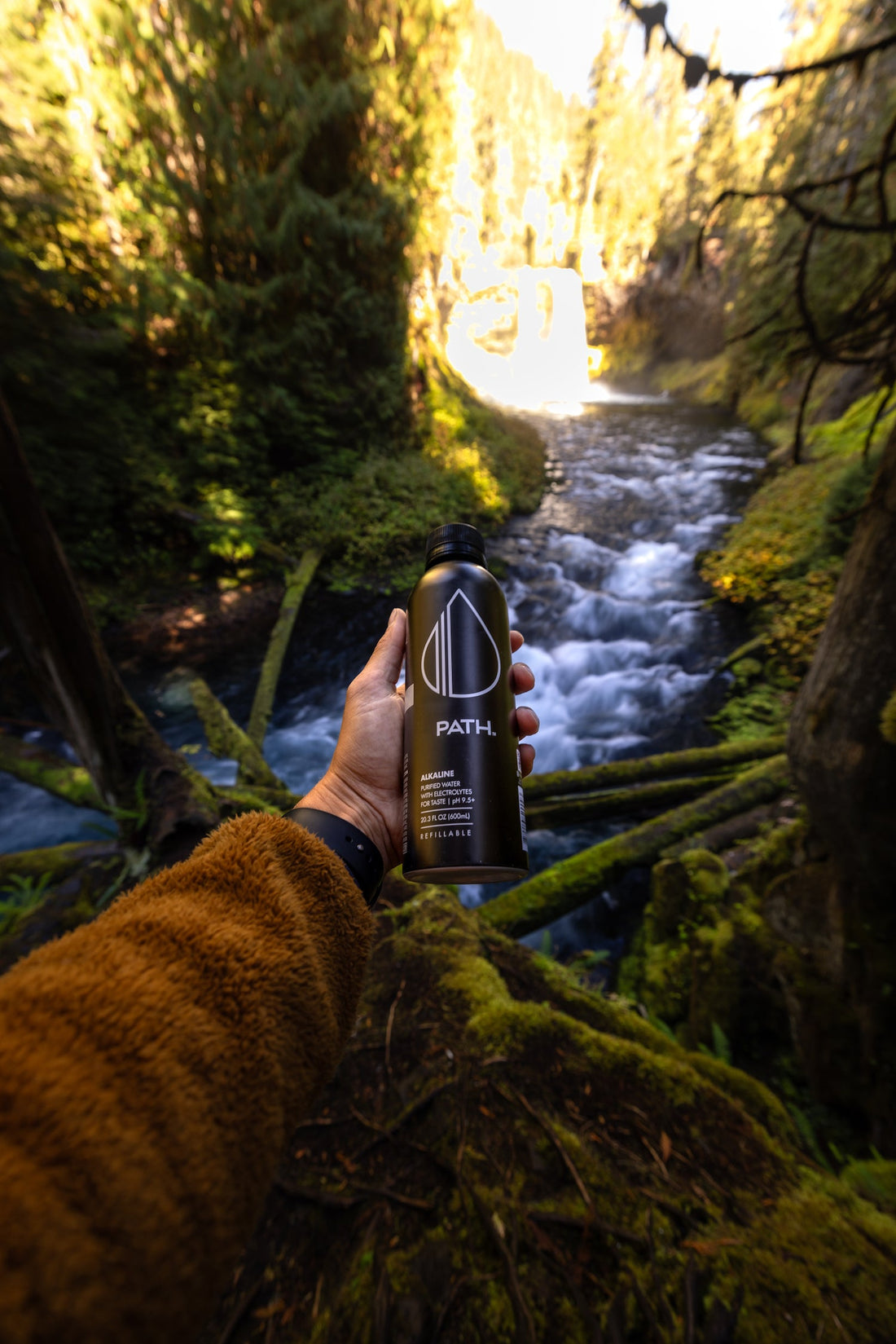Hand holding a PATH water bottle above a mossy forest stream with a waterfall in the background