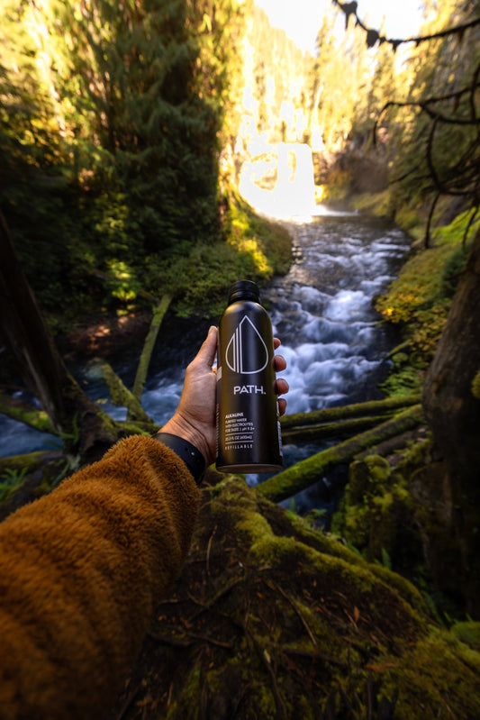 Hand holding a PATH water bottle above a mossy forest stream with a waterfall in the background