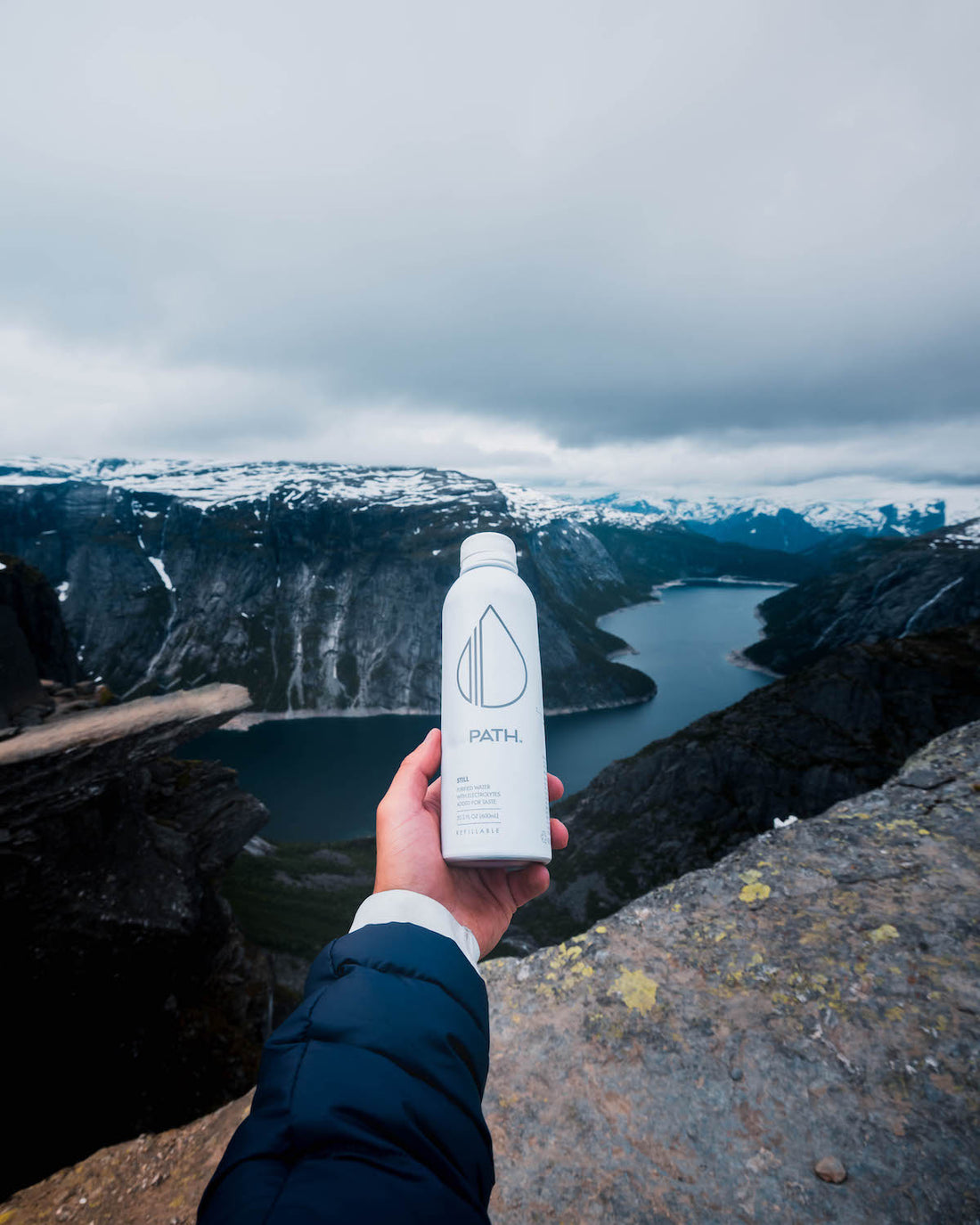 Hand holding a white PATH water bottle on a rocky cliff overlooking a snowy mountain fjord