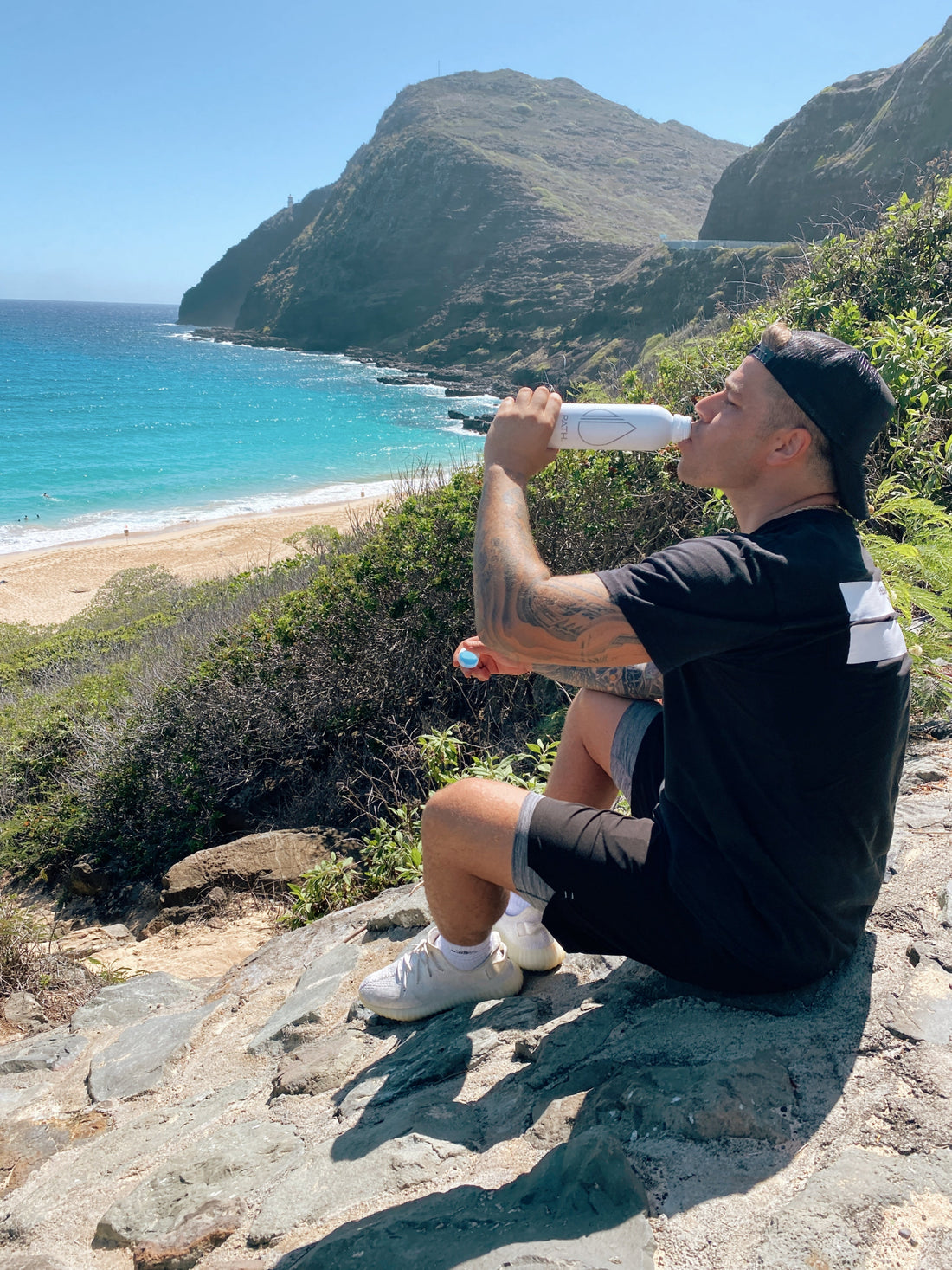 Man drinking from a white PATH water bottle while sitting on a rocky bluff above a turquoise ocean beach