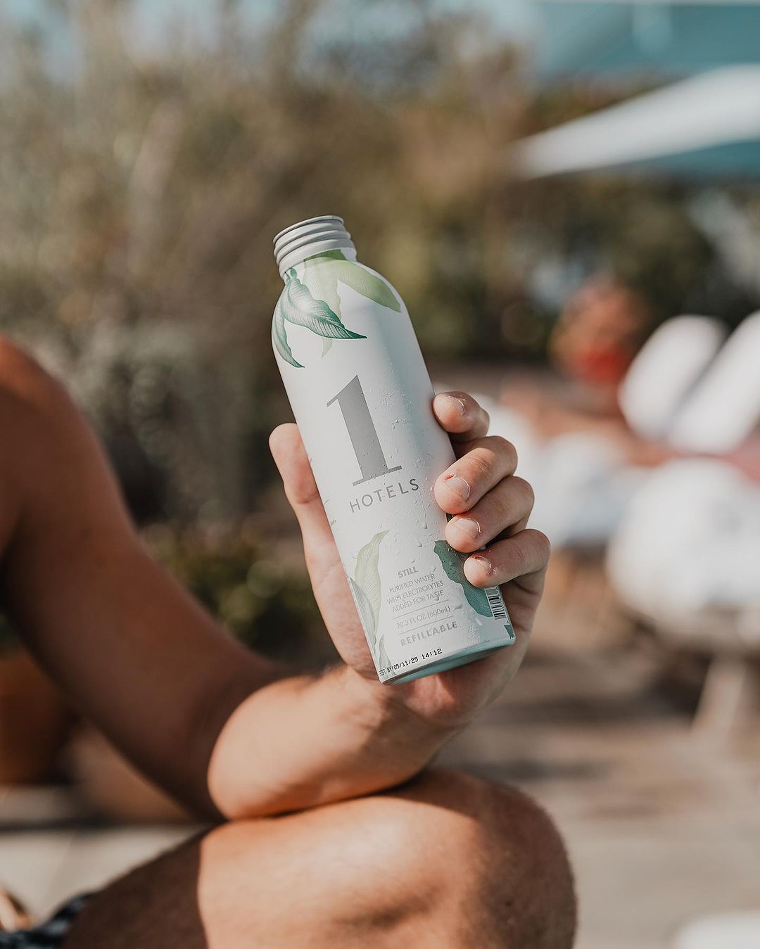 Hand holding a 1 Hotels refillable bottle showing 1 HOTELS logo and botanical design with blurred poolside background