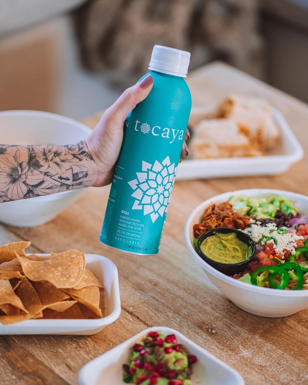 Tattooed hand holding a turquoise Tocaya water bottle above a table with tortilla chips and a rice bowl