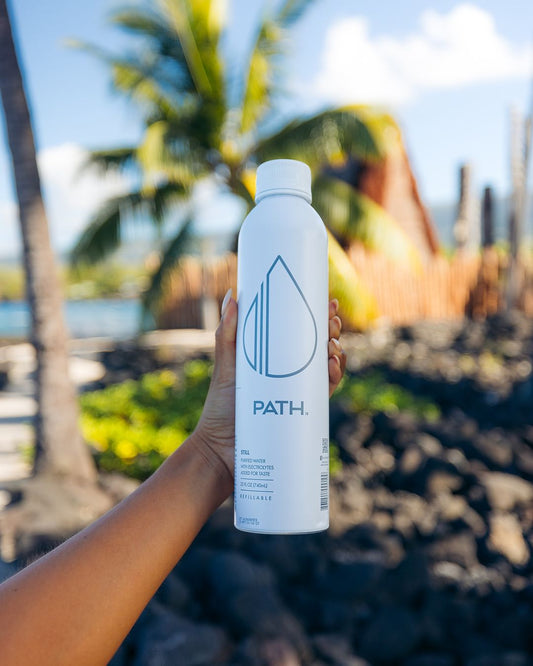 Front view of PATH white refillable water bottle showing logo and 'refillable' label held against blurred palm trees and rocks background
