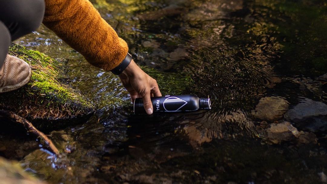 Hand placing a black PATH aluminum bottle into a shallow forest stream to refill