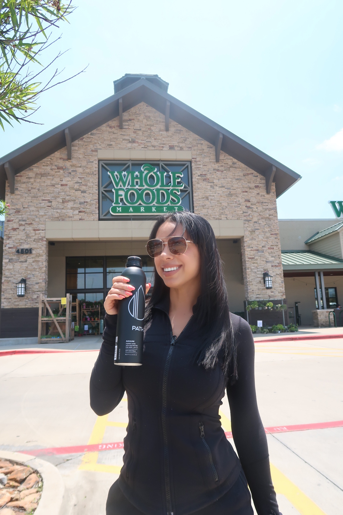 Smiling woman holding a black PATH water bottle outside a Whole Foods Market entrance