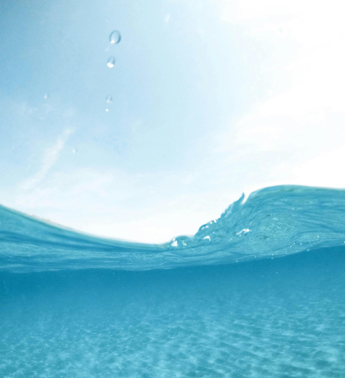 Split level view of clear blue ocean water with a small cresting wave and falling droplets, sunlit sandy seafloor