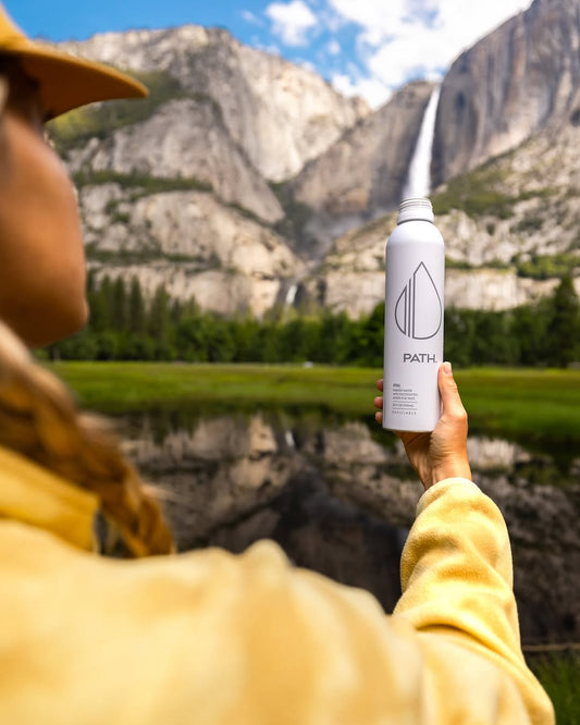 Person in a yellow jacket holding a white PATH aluminum water bottle toward a mountain waterfall and meadow reflected in a pond