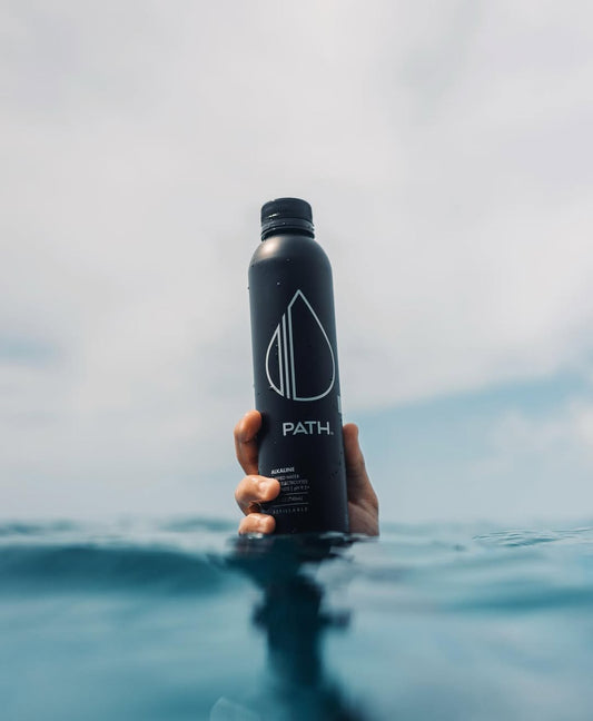 Hand holding black PATH water bottle above ocean surface with cloudy sky background