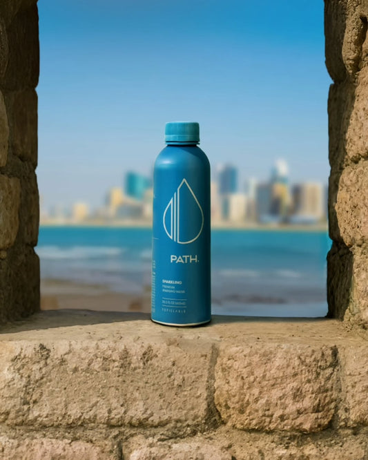 Front view of blue PATH refillable aluminum bottle showing logo and matte finish on a stone ledge with ocean and blurred city skyline background