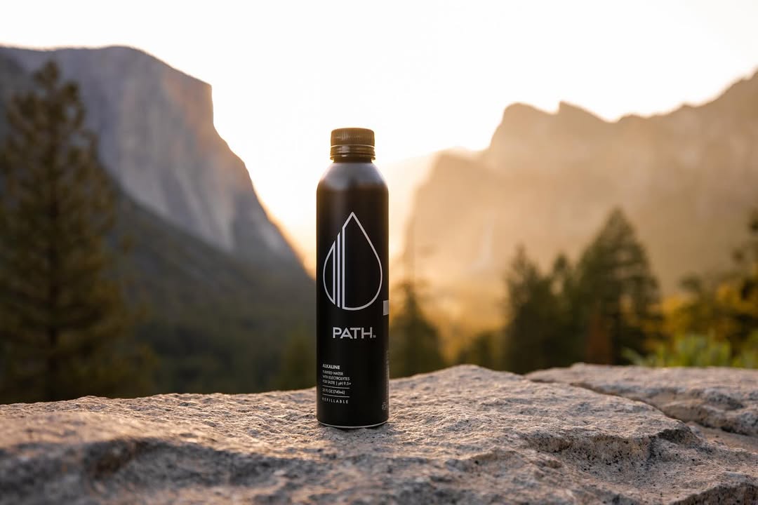 Front view of PATH aluminum water bottle showcasing white logo and matte black finish on a rock with mountains at sunrise