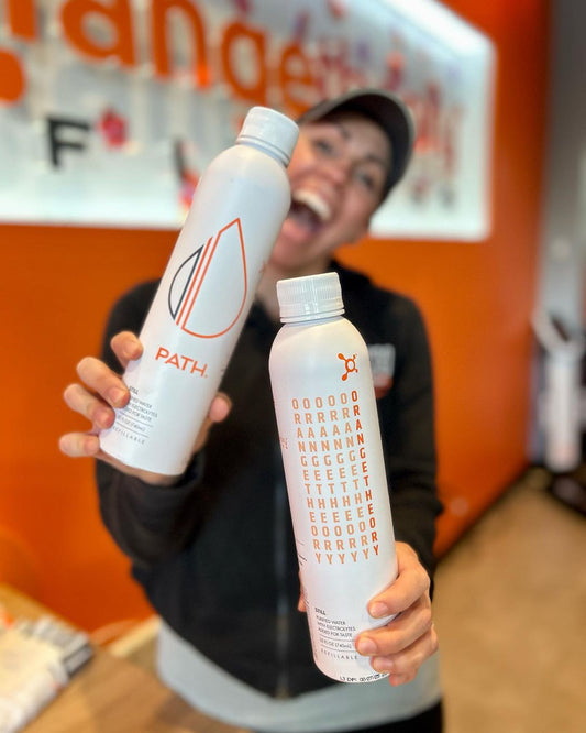 Smiling person holding two white PATH refillable bottles toward camera against orange gym background