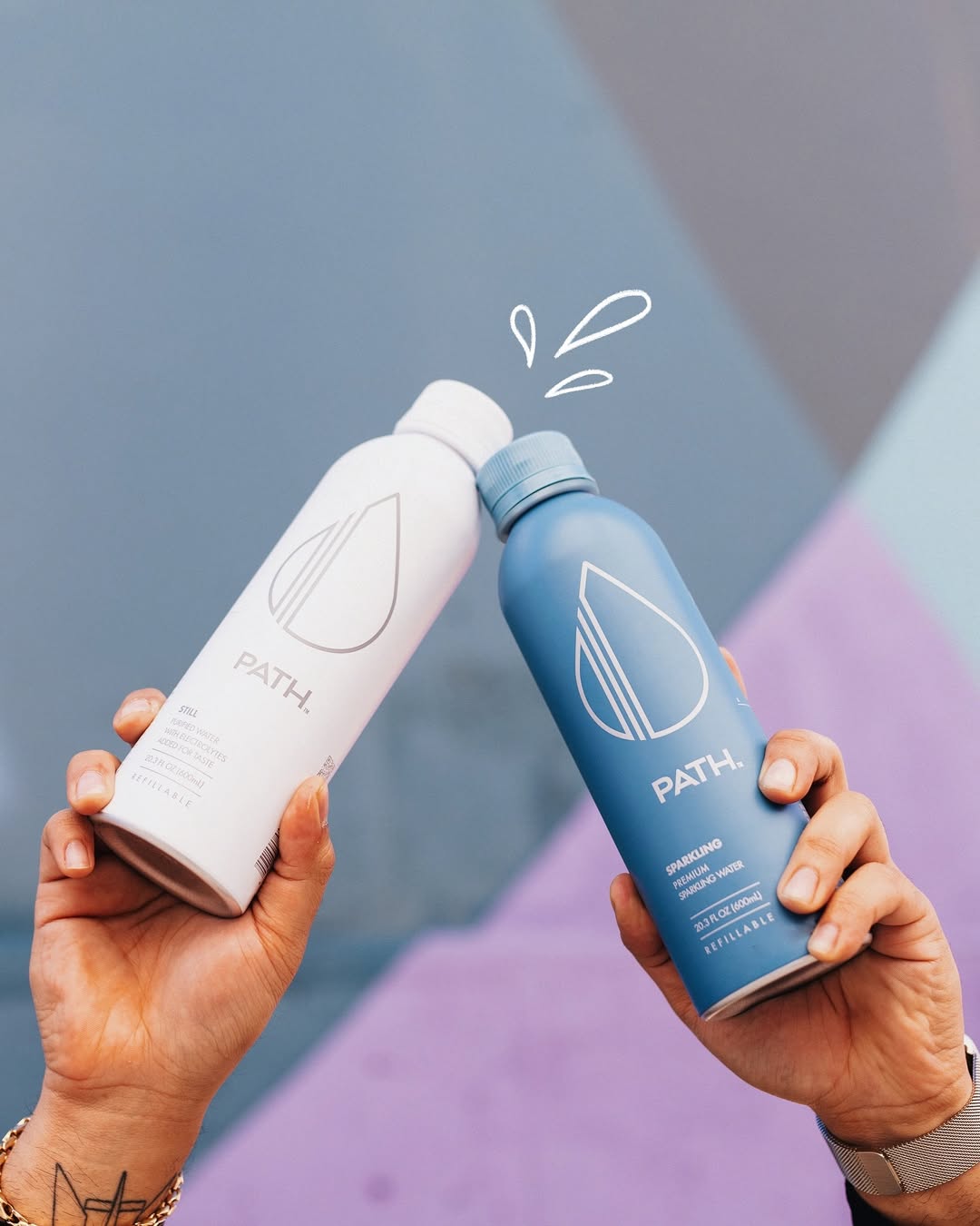Two hands clinking a white PATH refillable aluminum bottle and a blue PATH refillable bottle against a pastel geometric background
