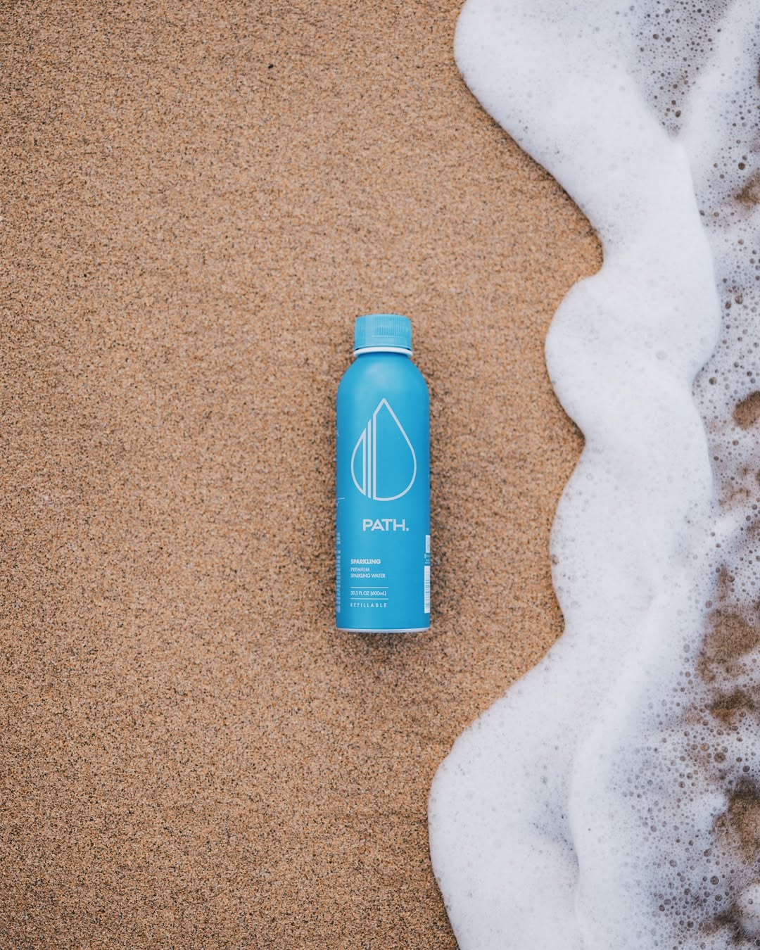 Top-down view of blue PATH aluminum bottled water showcasing PATH logo and refillable aluminum design on sandy beach with ocean foam