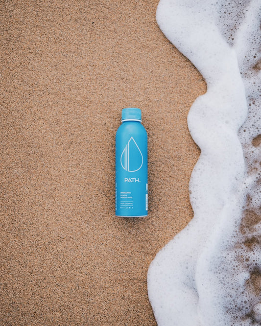 Top-down view of blue PATH aluminum bottled water showcasing PATH logo and refillable aluminum design on sandy beach with ocean foam