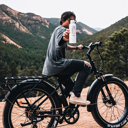 Person on a bicycle extending a white PATH water bottle toward camera on a mountain dirt road