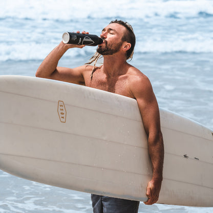 Surfer drinking PATH alkaline water from a black aluminum bottle while carrying a white surfboard at the ocean shore