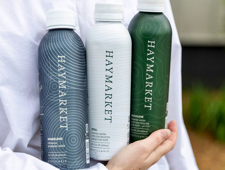 Close-up of three custom-printed aluminum bottled water bottles showcasing Haymarket designs in a person’s hands