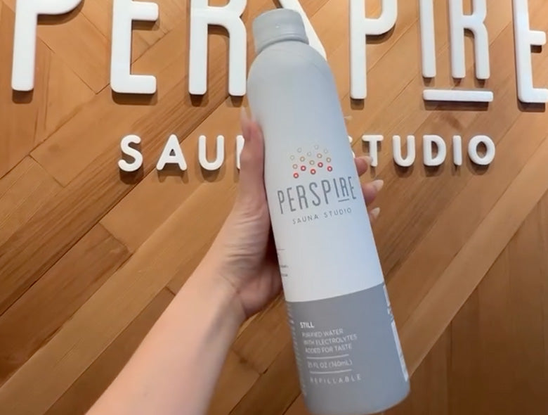 Close-up view of a custom aluminum bottled water bottle showcasing Perspire Sauna Studio branding with a wood-paneled background