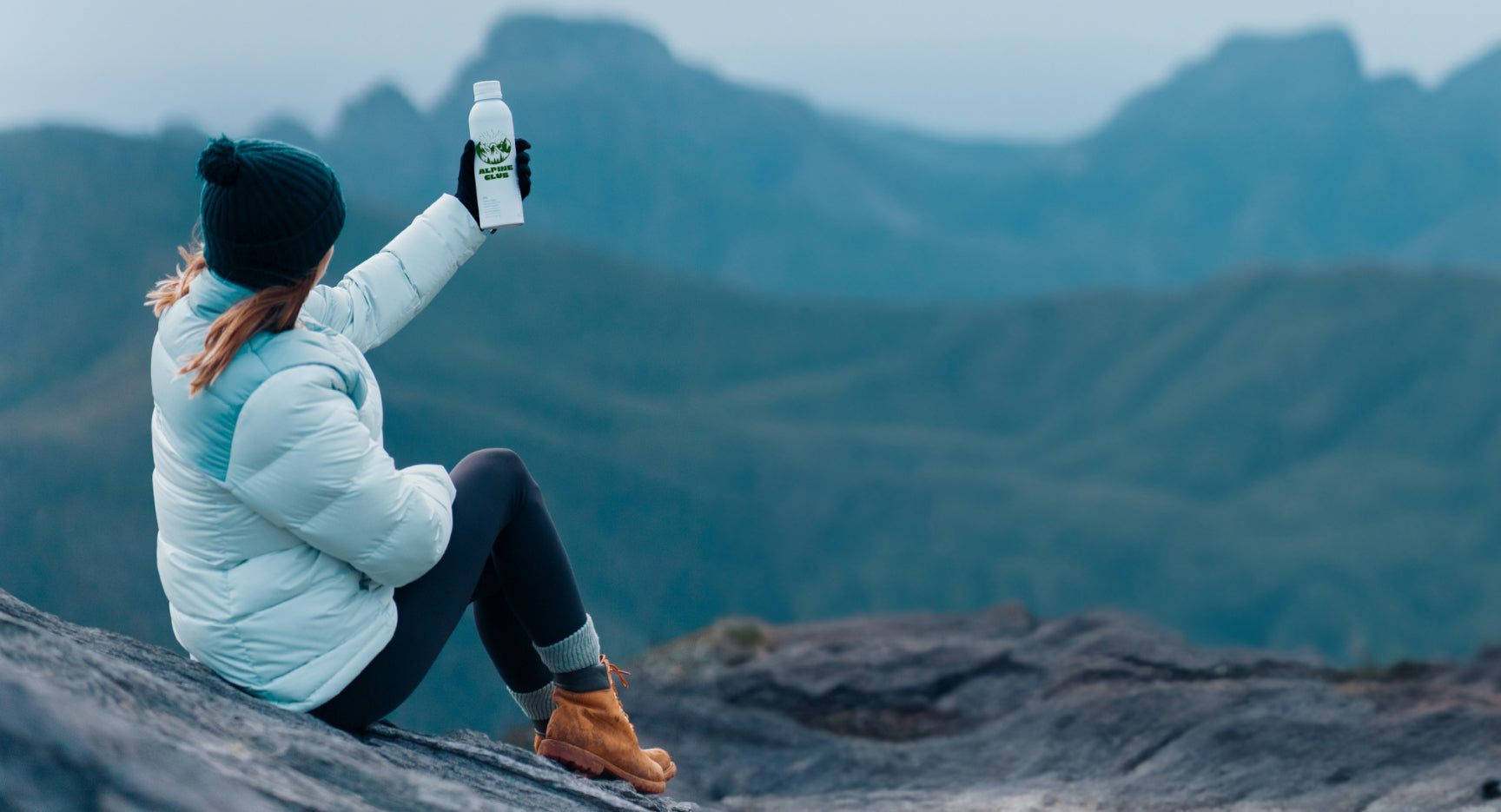 Woman seated on a rocky mountain holding a white bottle against a misty mountain landscape